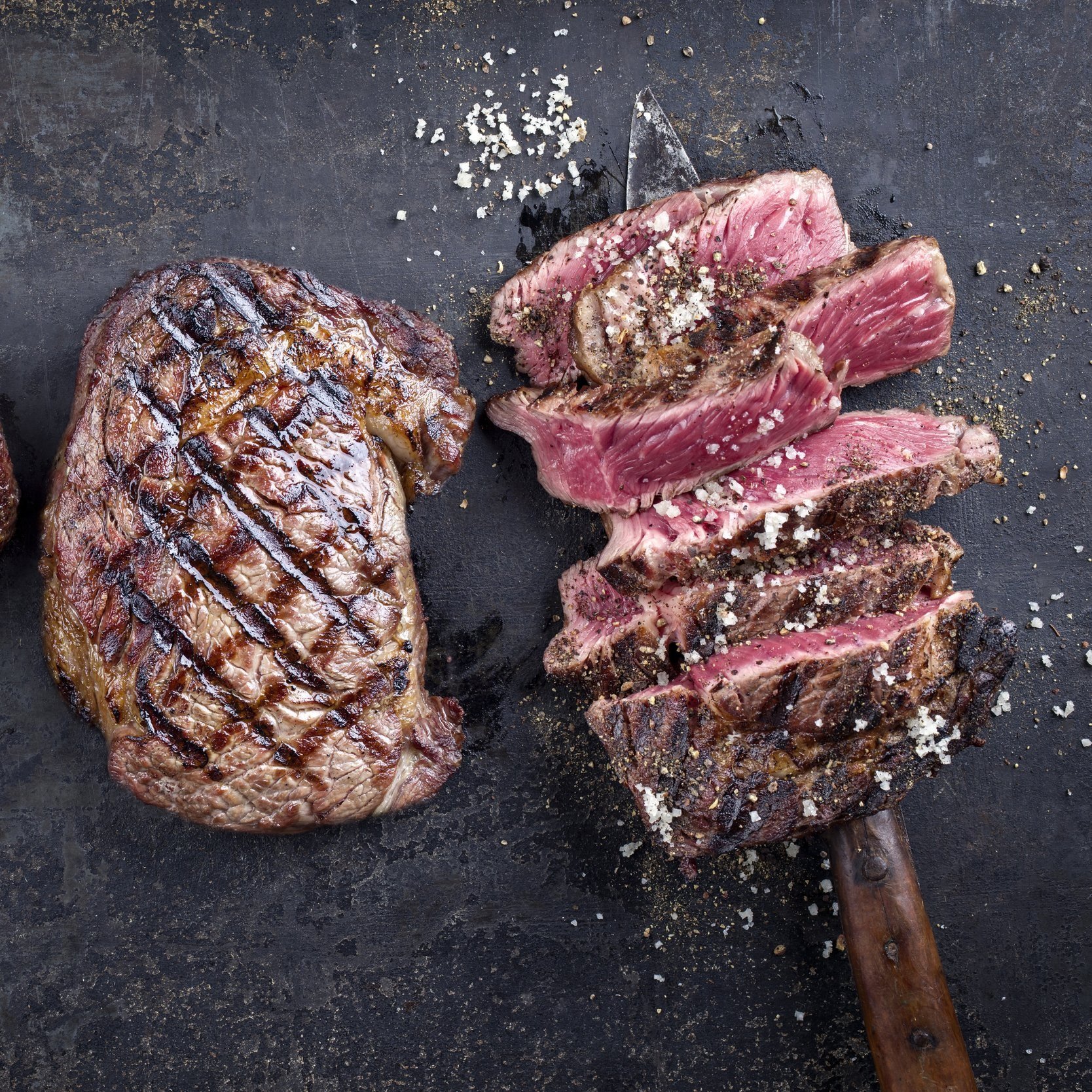 Grilled beef steak, perfectly seared and sliced, showcasing juicy pink interior and seasoning with coarse salt on a dark textured surface. Ideal for meat lovers and culinary enthusiasts.
