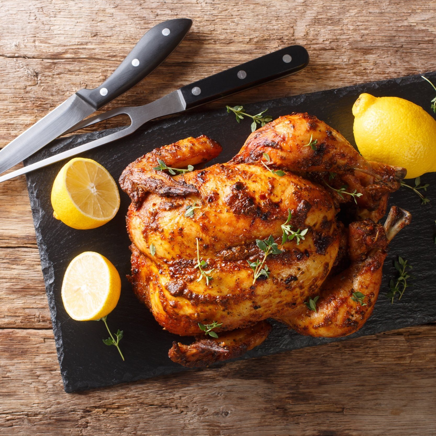 Perfectly roasted whole chicken garnished with fresh thyme and served with lemon slices on a black slate platter, alongside sharp kitchen knives on a rustic wooden background.