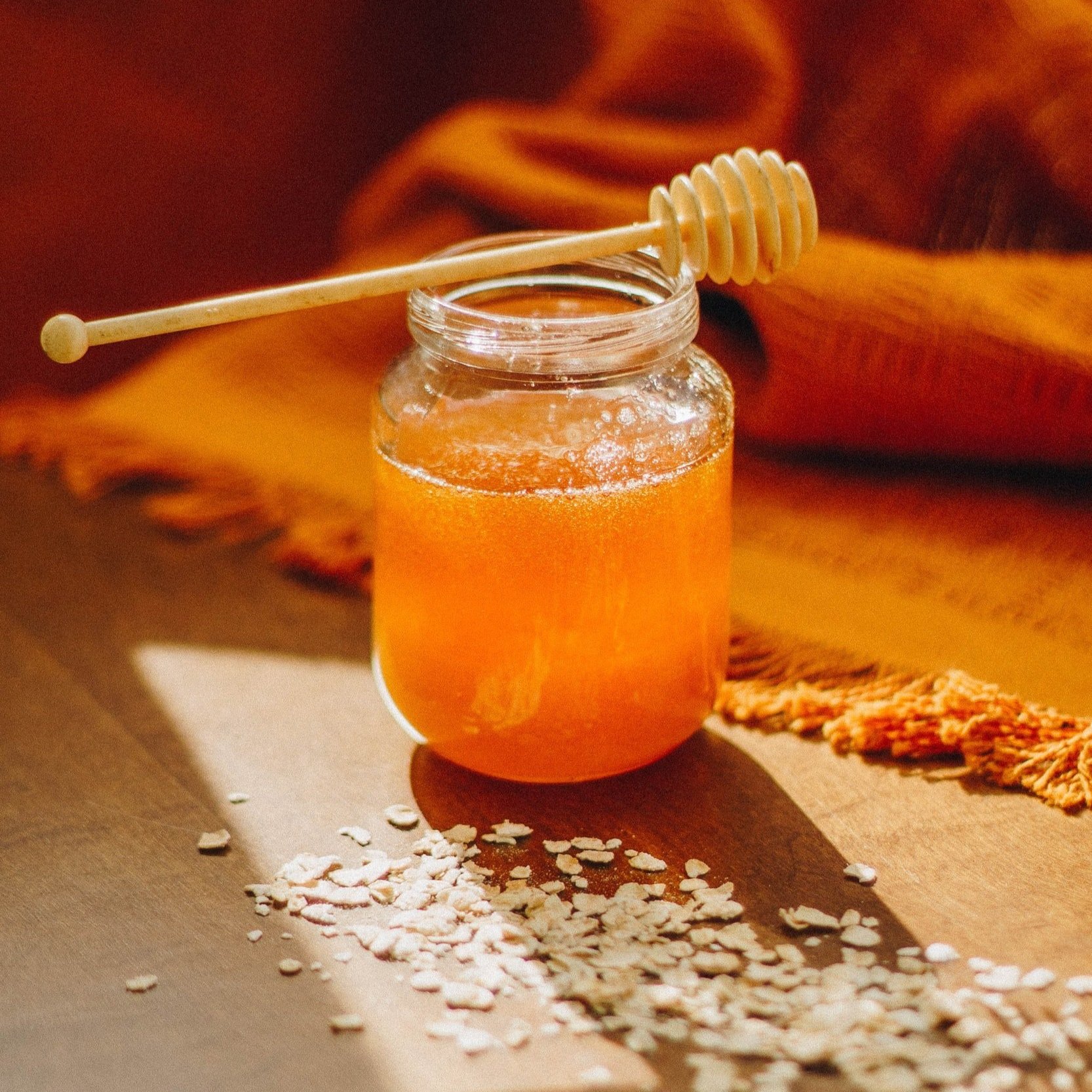 Glass jar filled with golden honey, accompanied by a honey dipper, set against a warm, textured background with scattered oats on a wooden surface.